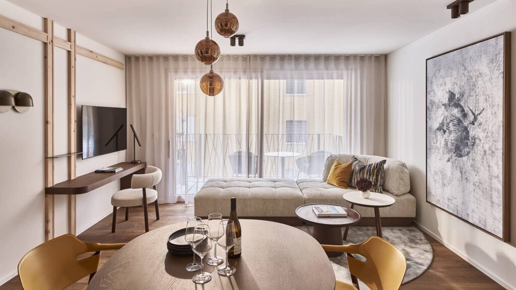 Wohn- und Essbereich eines modernen Apartments mit rundem Holztisch, gelben Stühlen, Sofa und Balkon mit Tageslicht